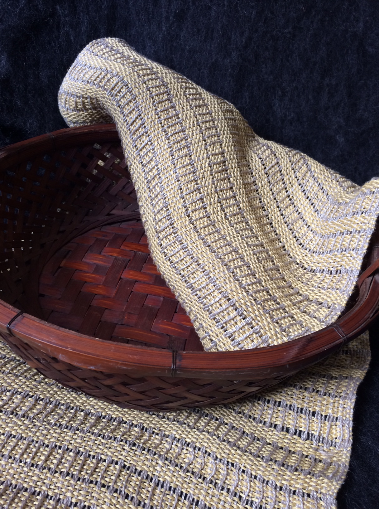 An oval rosy wood open weave basket rests on top of a 14 x 65 inch natural colored  linen table runner. The far end of the runner is folded back into the basket. The weave is made up of horizontal rows of  a natural color ladder stripes against a pale yellow background.